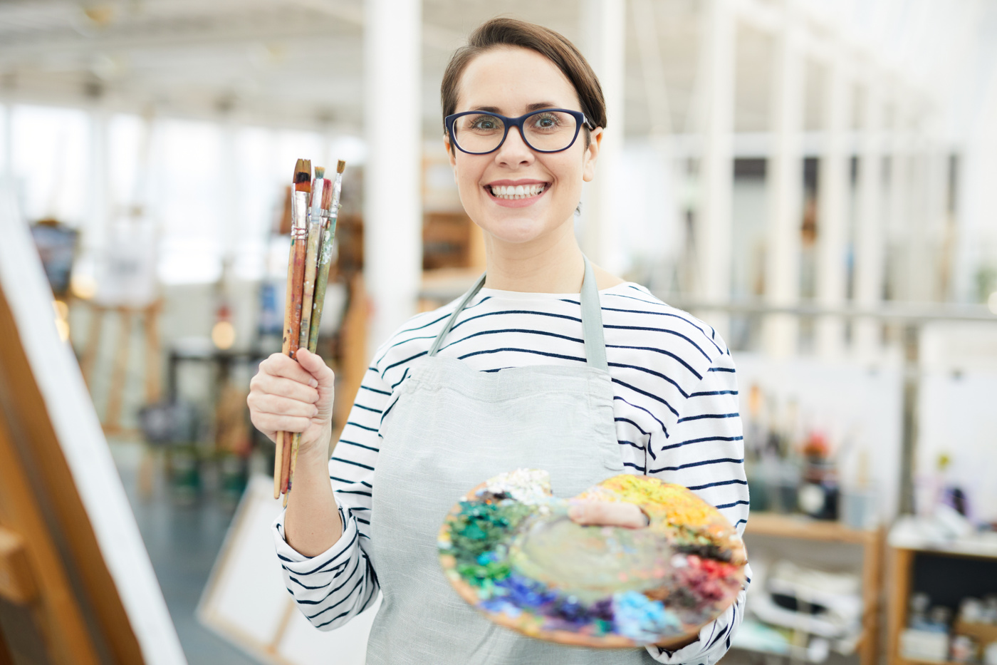 Eine Kunsttherapeutin hält Pinsel und eine Malpalette in der Hand und lächelt in die Kamera.