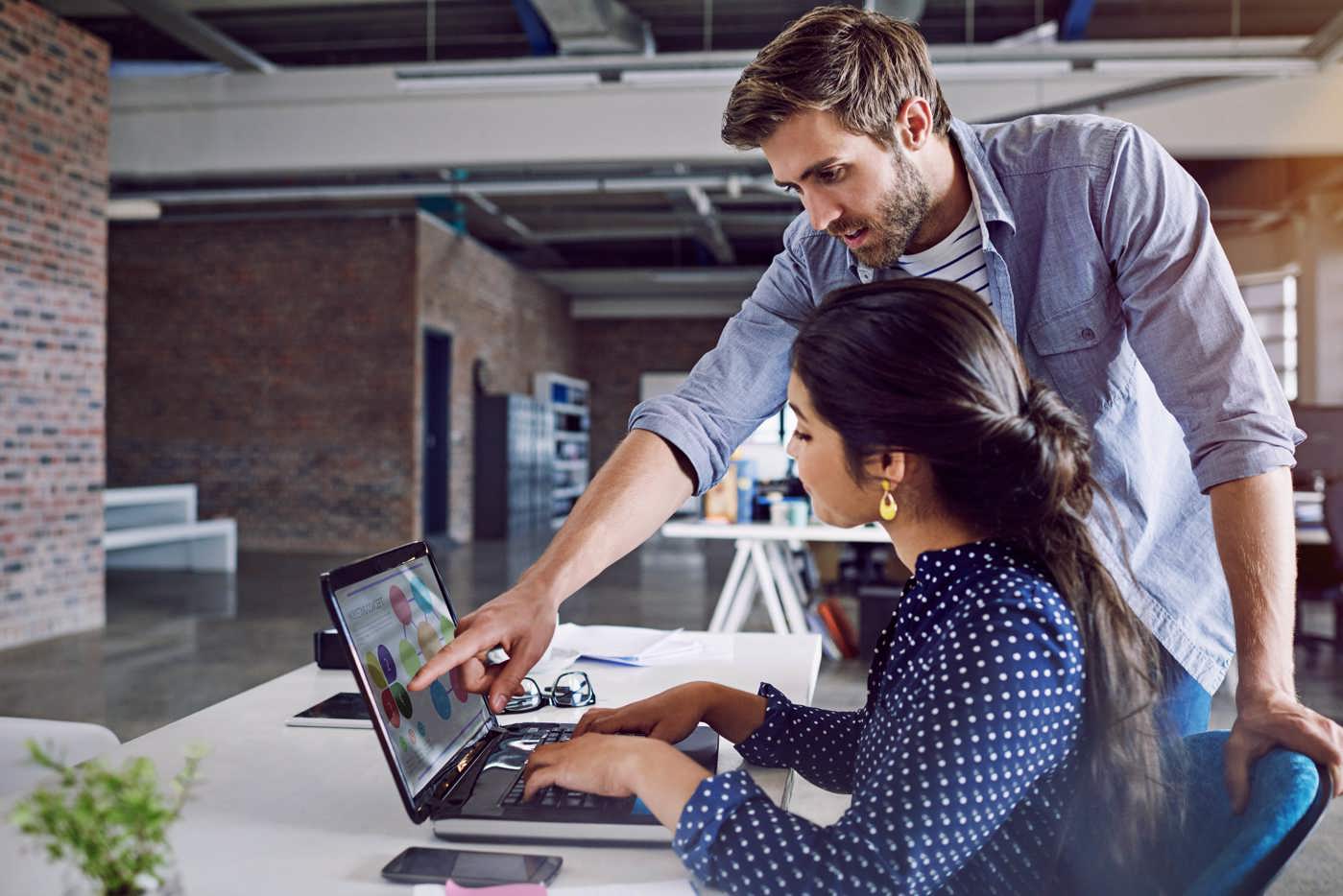 Ein Mitarbeiter erklärt einer Sachbearbeiterin Marketing und Verkauf das Marketing-Konzept auf dem Laptop.